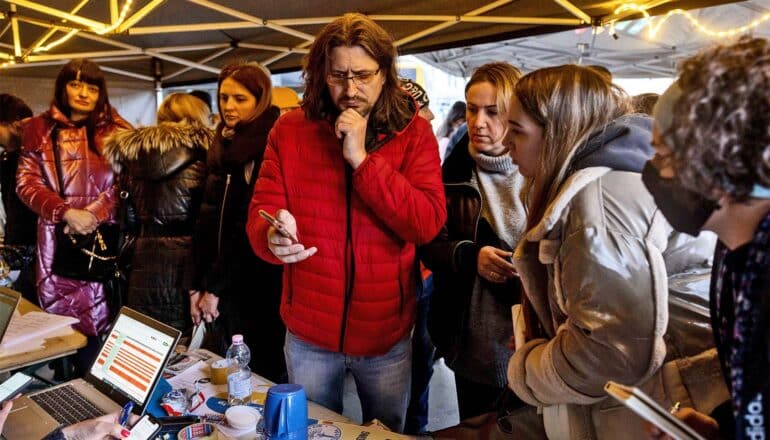 Refugees stand under a tent at a desk where aid workers help search for accommodations.