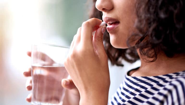 A teen girl takes a pill while holding a glass of water.