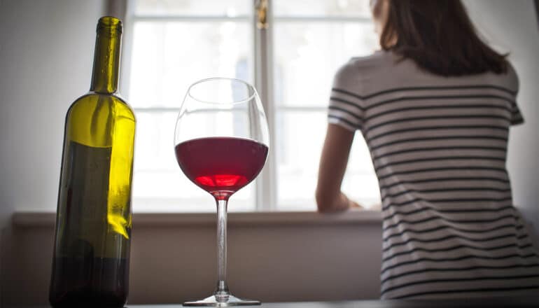 A woman looks out the window as a wine bottle and glass of wine stand on the table behind her.