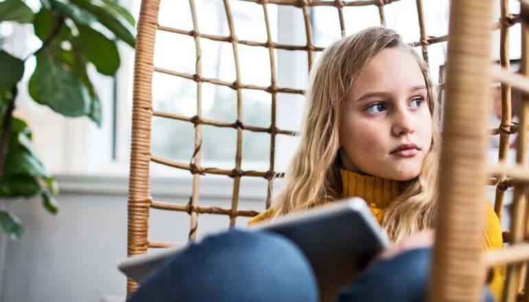 A young girl sitting in a chair with a tablet computer looks off into the distance.