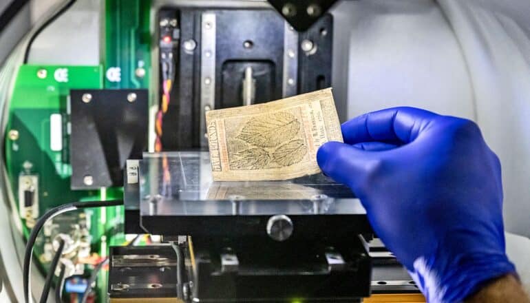 A researcher wearing a blue gloves places Ben Franklin's money under a microscope.