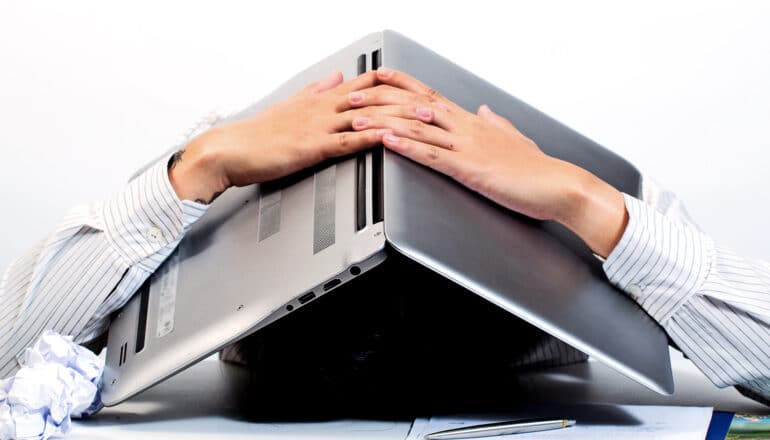 A person holds their laptop over their head as they lay on their desk in frustration.