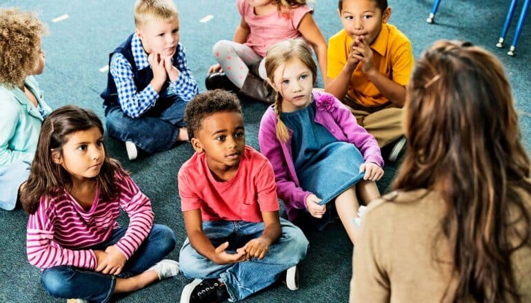 A group of young kids sit on a classroom carpet while listening to a teacher.