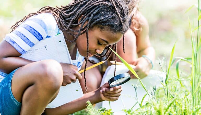 A girl with a magnifying glass looks down into some grass.