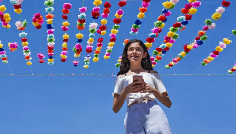 smiling Latinx teen holds phone beneath colorful bunting