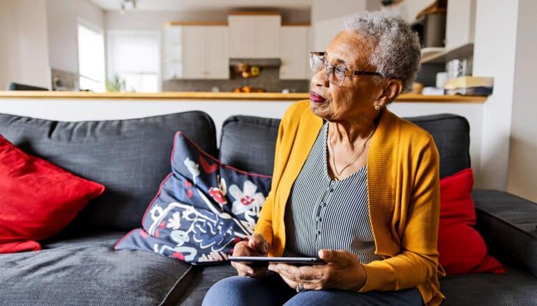 An older woman sits on her couch with a tablet computer in her hands.