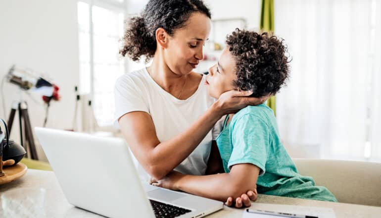 A mom hugs her child while sitting at a kitchen table in front of a laptop.