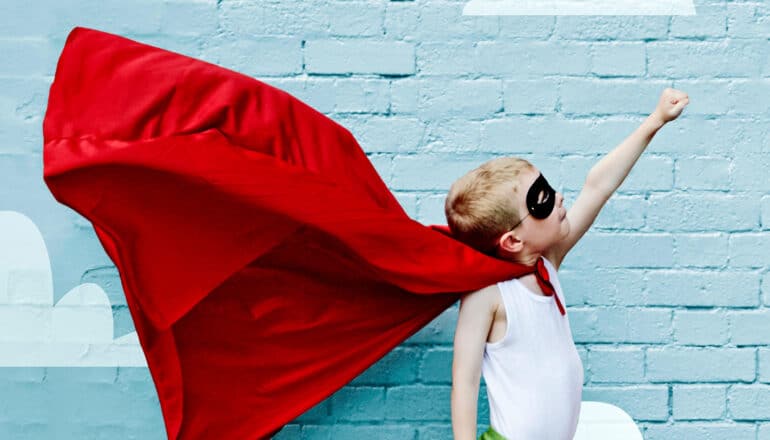 A young boy dressed up as a superhero with a red cape and black mask imitates flying against a blue brick wall.