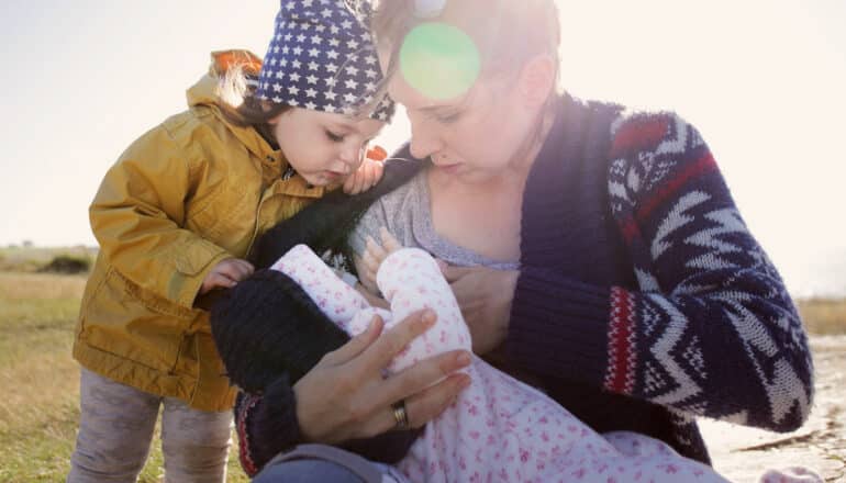 adult breastfeeds baby outdoors as toddler looks on