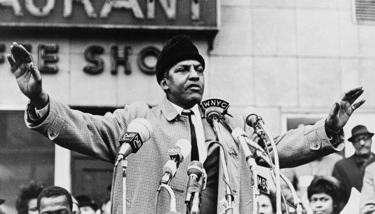 Bayard Rustin speaks into press microphones with arms spread wide