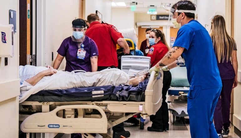 Hospital workers move patients through a hallway while wearing face masks and personal protective equipment.