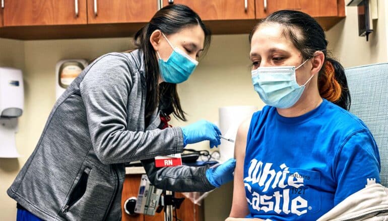 A young woman gets a COVID vaccine from a health worker in a blue mask.