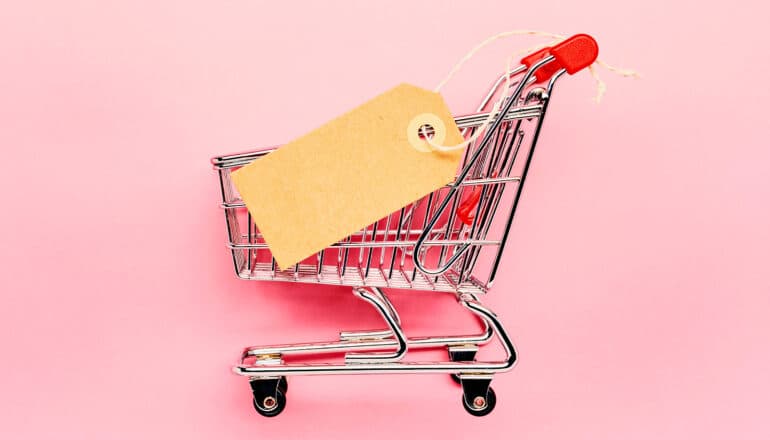 A shopping cart toy with a blank price tag on it against a pink background.