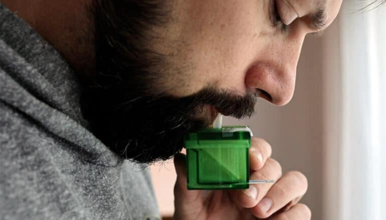 A bearded man blows into a small green, boxy device.