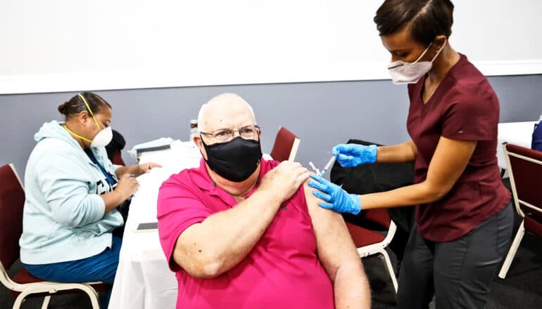 An older man gets a Covid vaccine from a health worker in scrubs.