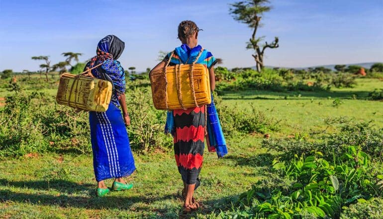 Two young women carry cans of water on their backs while walking on grass.