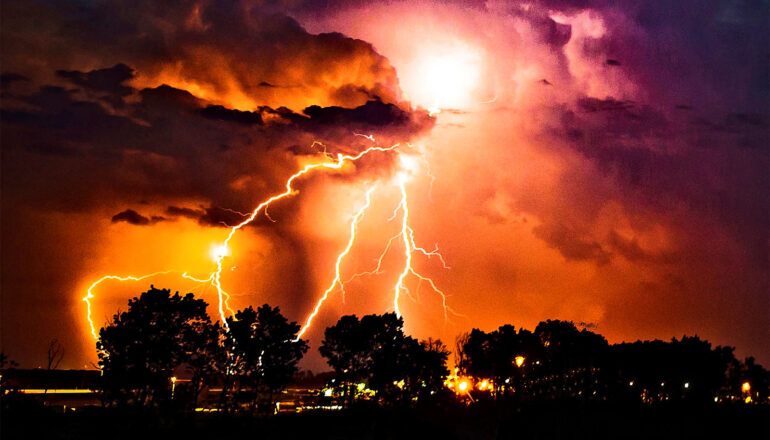 Lightning strikes over a row of trees in the foreground.