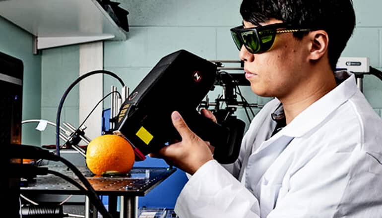 Shin points a large, black, handheld device at an orange resting on a surface while wearing protective goggles.