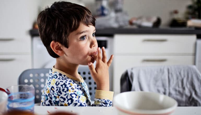 A young boy licks his finger while standing next to a dinner table.