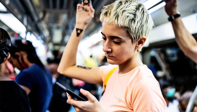 A woman on a subway train looks at her phone and holds a handle on the ceiling.