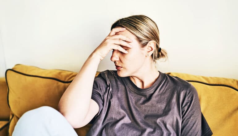 A woman puts her hand to her head while sitting on a couch.