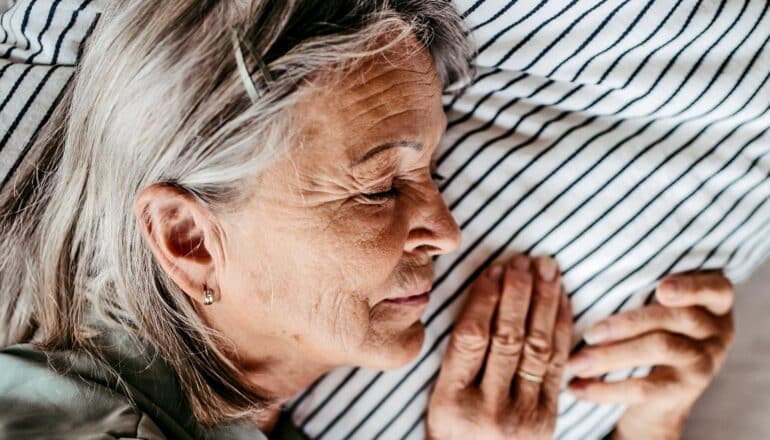 An older woman sleeps with her hand on the pillow next to her head.