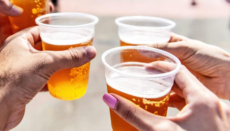 People hold up plastic cups of beer while outside.