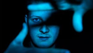 A man frames his face with his fingers while cast in blue light.