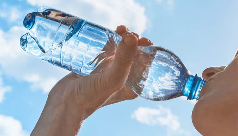 A person drinks a large bottle of water with blue sky in the background.