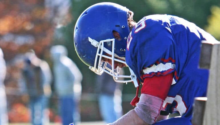 A college football player sits on a bench while looking down at the ground.