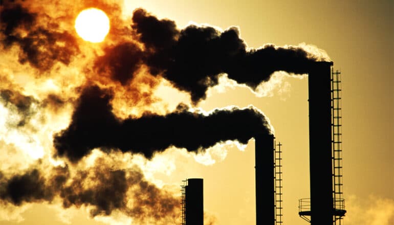 Black smoke pours from three smokestacks at sunset.