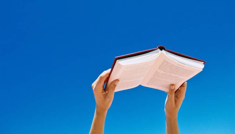 A person holds up a book against a deep blue sky.