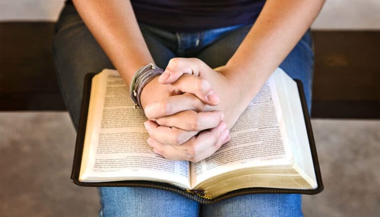 A young woman clasps her hands over an open Bible while seated.