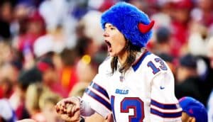 A Buffalo Bills fan wearing a blue fuzzy hat with red horns on it shouts for the team at a packed game.