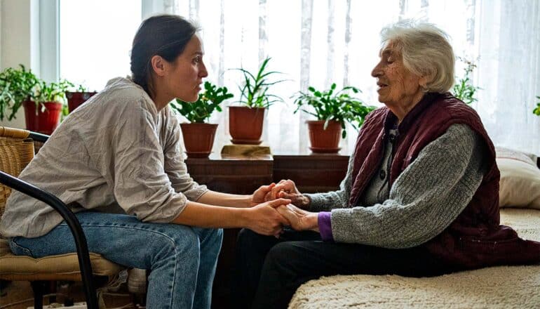 An elderly mother and younger daughter sit while holding hands and talking.