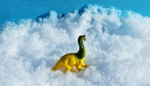 A yellow and green toy dinosaur sits on top of a pile of white snow.