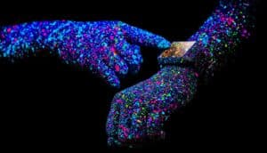 A person uses a smartwatch with their arms covered in specks of colorful paint against a black background.