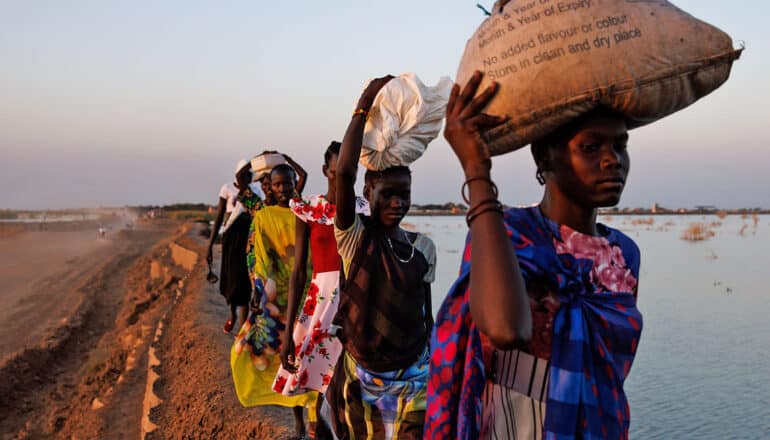 row of people carrying parcels on their heads walk at edge of water