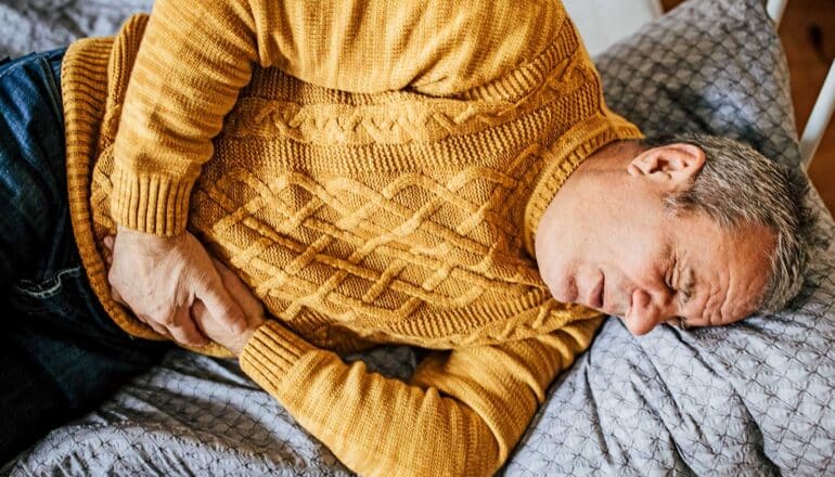 A man lays on a bed grimacing and holding his stomach.