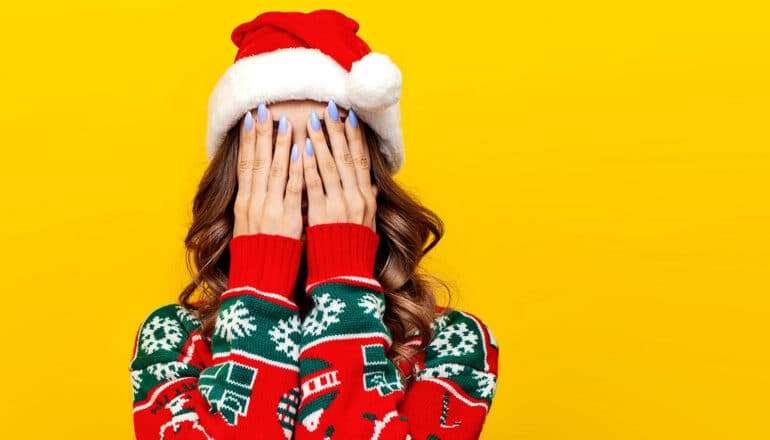 A woman wearing a holiday sweater and Santa hat puts her hands over her face.