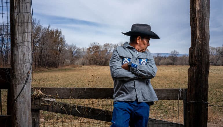 man in cowboy hat stands against fence, looks over his shoulder