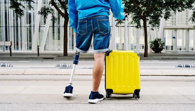 A man with his left leg amputated stands on a sidewalk with a rolling suitcase.