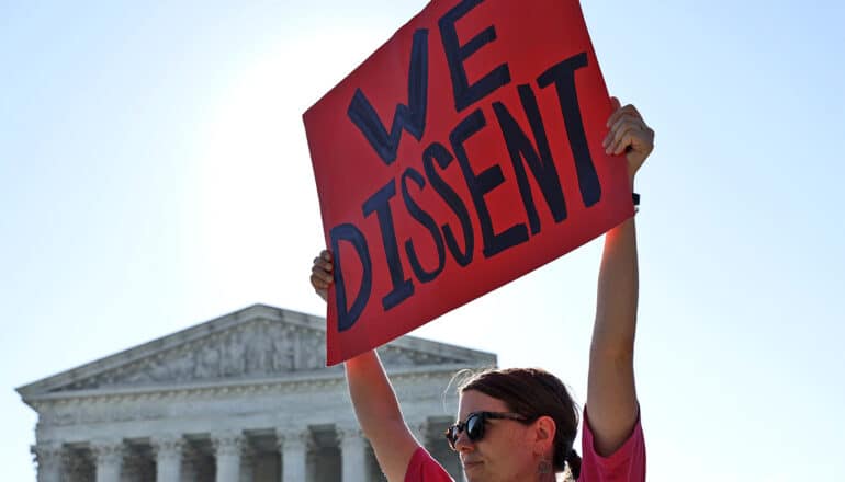 A woman holds a sign up in front of the Supreme Court building that reads "We dissent."