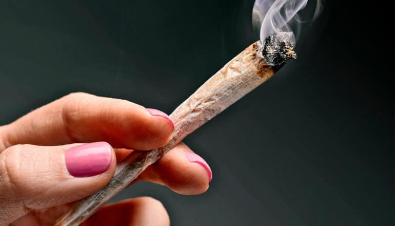 A woman with pink painted nails holds a joint with smoke coming out of it.