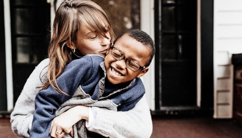 A mother holds her son and kisses his cheek as he laughs and pulls away slightly.