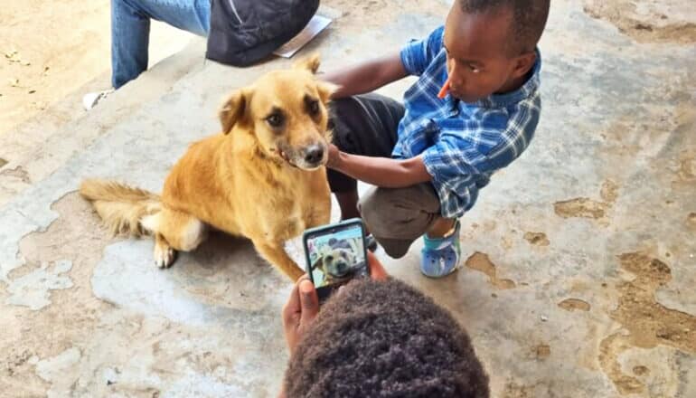 A person takes pictures of a dog with a smartphone while a young boy holds the dog still.