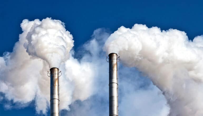 Two smokestacks releasing white smoke into the blue sky.