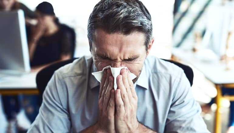 A man blows his nose while sitting in an office with people behind him.