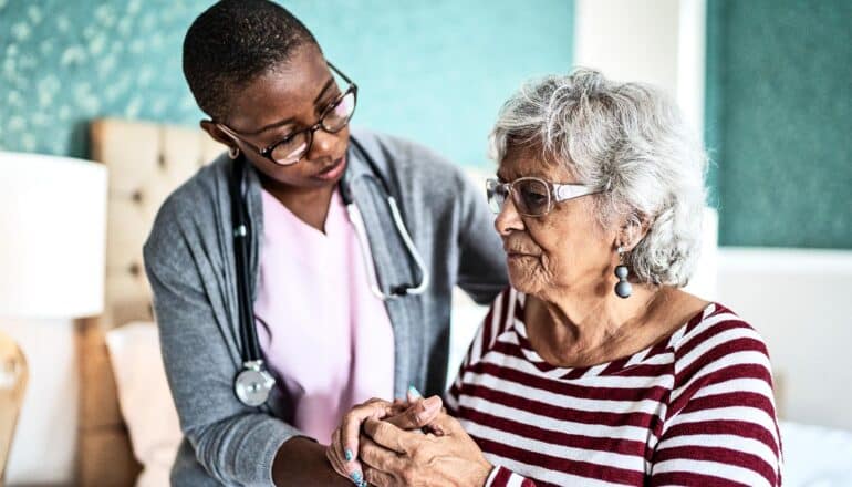 A nursing home health care worker speaks with an older woman in her room.