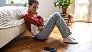 A teen girl sits on the floor at the foot of her bed holding her head with one hand and looking at her phone on the floor.